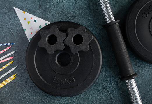 Heavy dumbbells barbell weight plates with cone shaped hat and colorful cake candles. Gym exercise equipment as a gift for birthday party. Healthy fitness lifestyle flat lay composition concept.