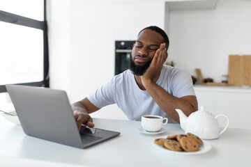 Fototapeta na wymiar Tired black man using laptop sitting at kitchen table
