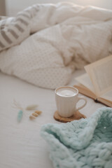 Fototapeta na wymiar Cup of coffee with milk and a book on the bed with white linens and blanket. Morning coffee in bed concept.