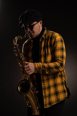 Obraz na płótnie Canvas Man Saxophonist in Yellow Checkered Shirt, Cap and Stylish Sunglasses Holding an Alto Saxophone. Black Background. Close-up Portrait. High quality photo