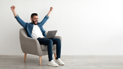 Overjoyed Arab man sitting in armchair with laptop, celebrating online win or work success against white studio wall