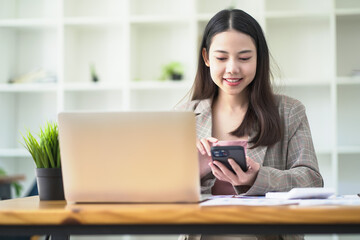 Asian woman smiling and looking at mobile phone in hand while working online on laptop.