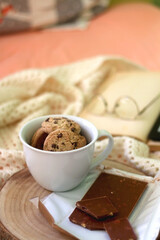 Mug of chocolate chip cookies, bar of chocolate, open book, reading glasses and e-reader on a bed. Selective focus.