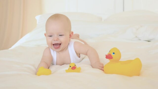 A happy laughing child is lying on his parents' bed after a bath or shower playing with rubber ducks. Clean dry baby in the bedroom. Bathing and washing small children. Hygiene of children.