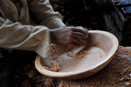 Antebrazos y manos movidas lijando el interior de un plato de madera en foco con restos de madera en su interior y en la mesa.