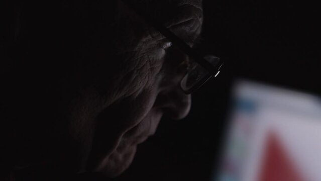 Senior citizen wearing glasses watching data on computer monitor sitting at table in dark room