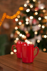 Obraz na płótnie Canvas Two red mugs on wooden table in the living room with christmas lights bokeh. Drinking hot beverages on holiday seasson at cozy apartment home. Chalet winter cottage core lifestyle.