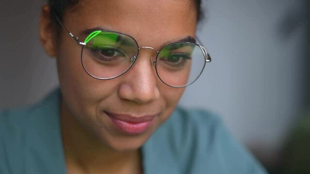 Smiling ethnic female student in eyeglasses using computer, flicker and reflections on the lenses, web surfing, researching topic, woman working online, watching movie, close-up