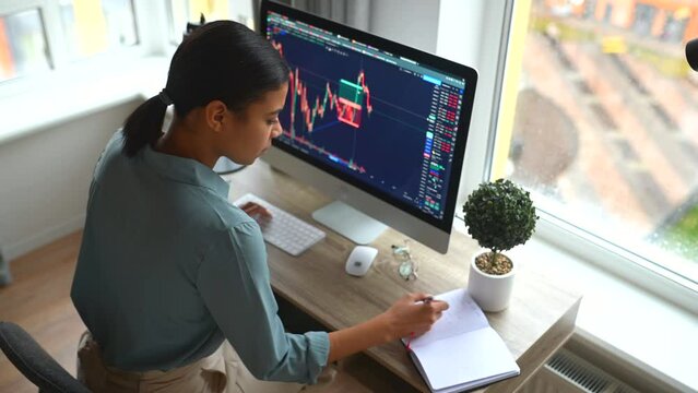 Smart and concentrated multiracial female freelancer working remotely from home, analyzes data of trading graph of stock market, develops business strategy, takes notes