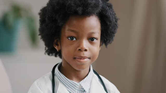 Cute serious african american kid girl in medical clothes dressed in white coat standing indoors posing looking at camera playing pretending to be doctor future profession concept close-up portrait