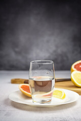 Vertical front view on citrus water in glass on white plate on light table against dark gray background. Sliced lemon and grapefruit on wooden board. Selective focus. Copy space.