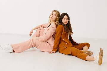 Modern young women in fashionable pastel suits with unbuttoned blazer sitting on a white background.