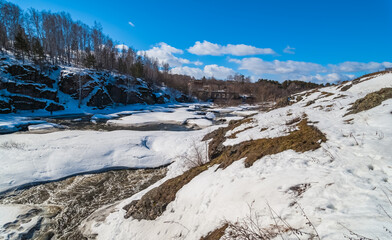 A spring landscape with a river, rocks, ice, snow, trees, a ruined brick building and a blue sky with white clouds