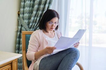 Serious mature woman reading papers documents sitting in armchair at home.