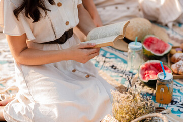 cropped photo of young woman in dress reading book while enjoying picnic in summer park. Sitting on blanket. Croissants, watermelon and cold summer drink