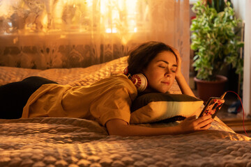 Relaxing young woman in headphones is lying on the bed with a pillow, and looking at her...