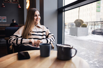 Beautiful smiling calm young woman drinking coffee looking out the window while sitting at a table in a restaurant. Morning coffee