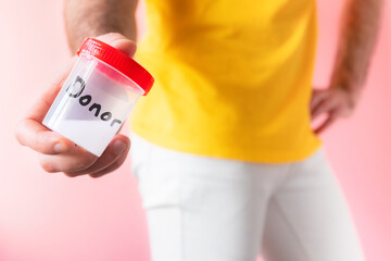 A man in white jeans holds a plastic jar with sperm sample with text donor in his hands. Close up. Pink background. The concept of artificial insemination and sperm donation