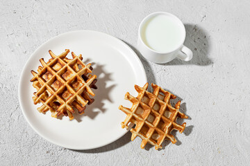 Freshly baked Belgian waffles on a white plate and a cup of milk on a light table, tasty breakfast...