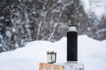 A black matte thermos and a mug with a spruce twig stand on a snow-covered stump.Warm tea in the winter frost.Minimalistic subject shot