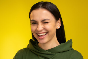 Cheerful caucasian brunette woman looking at camera and winking while showing her stylish clothes. Studio shot, isolated on yellow background