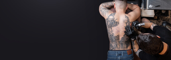 Professional tattoo artist makes a tattoo on on male back on a dark background with copy space for...