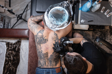 Professional tattoo master with a tattoo machine stuffs a knight's drawing on a man's back, top...