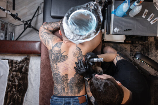 Professional tattoo master with a tattoo machine stuffs a knight's drawing on a man's back, top view. Workflow tattoo artists