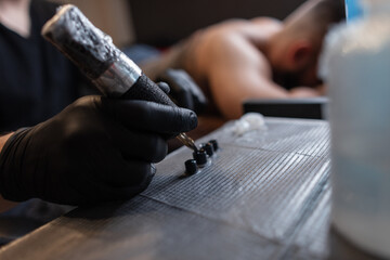 Man tattoo master professional in black gloves with tattoo machine picking up ink, workflow. Male hand close up