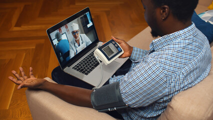 High angle view of African-American man with blood pressure meter consulting doctor online on laptop. Sick guy measuring blood pressure and consulting cardiology on computer at home - obrazy, fototapety, plakaty