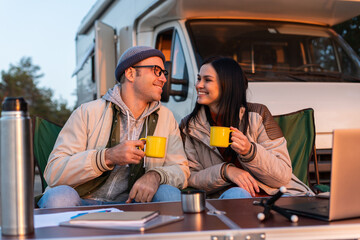 Smiling man holding cup with tea and squints against the bright sun near his woman. Happy family sitting at the nature and drinking hot tea from cups at camp fire in cozy forest