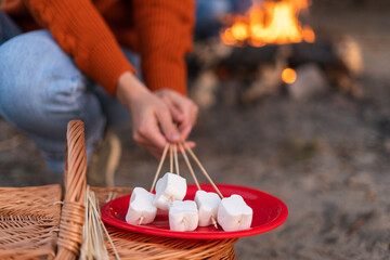 Cropped view of the caucasian woman enjoying outdoor recreation while holding marshmallows at the sticks and preparing roasted it over the fire with her boyfriend. Stock photo