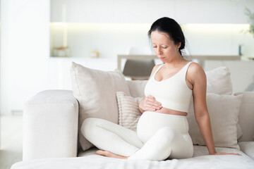 An attractive pregnant woman is sitting on a sofa in the living room of her home. She keeps her hand on her stomach so she can feel her baby move. Last months of pregnancy.