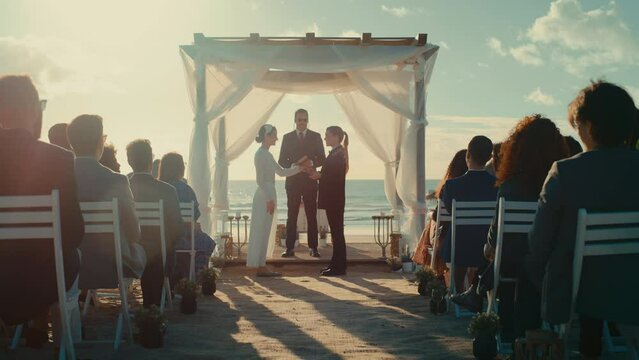 Beautiful Female Queer Couple Exchange Rings and Kiss at Outdoors Wedding Ceremony Near Ocean. Two Lesbian Women in Love Share Their Big Day with Diverse Multiethnic Friends. LGBTQ Relationship Goals.