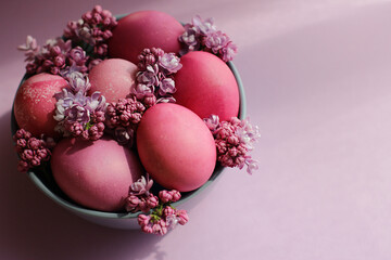 A grey bowl with bright pink Easter eggs and lilac flowers on a purple background. Easter decor. Selective focus