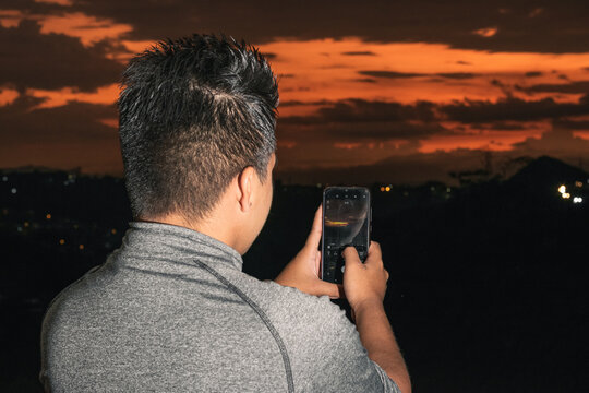 young latino man with brown skin, taking a picture with his cell phone of a beautiful sunset in the amazing south american mountains. student taking a picture for his thesis. education concept.