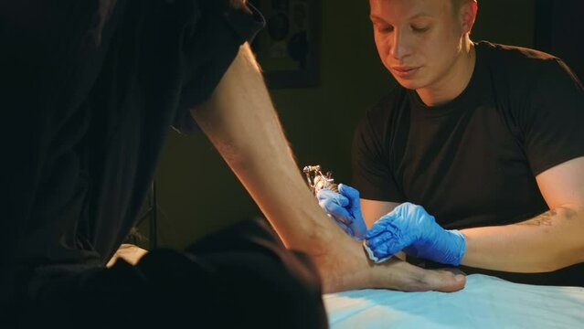 Close-up face of young adult focused male tattooist, tattoo artist works with customers shaved foot. Master tattooer man in medical hand gloves with old school coil needle machine fills clients leg.