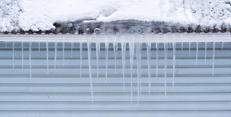 Several icicles hang from the roof against a gray plastic siding wall. Cloudy winter day. Background.