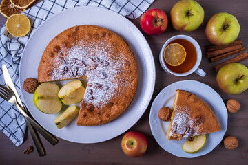 Top view close-up of sliced apple pie with walnuts and ripe apples. Piece of cake on plate and tea with lemon. Homemade cakes with seasonal fruits. Selective focus.