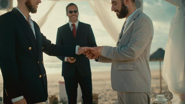 Close Up of Handsome Gay Couple Exchange Rings and Kiss at Outdoors Wedding Ceremony Venue Near the Sea. Two Happy Men in Love Share Their Vows and Get Married. LGBTQ Relationship Goals.