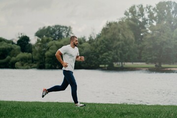 Sporty European man dressed in t shirt, trousers and sneakers and runs along river, trains...