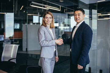 Fototapeta na wymiar Asian businessman shakes hands with business woman employee, happy team of workers looking at camera and smiling, successful conclusion of negotiations