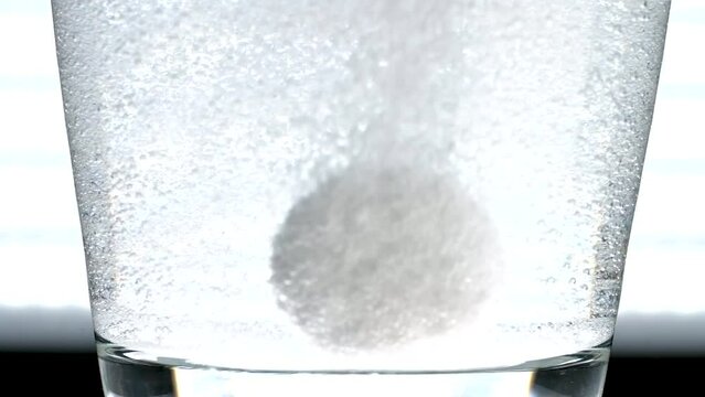 Antacid tablets dissolving in a backlit glass of water before floating up out of frame.  Dense bubbles stick to the inside of the glass, macro view of carbonation release from otc medication.