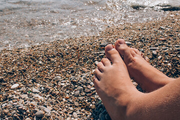 Unrecognisable child sitting on the beach near water, children's legs against sea shore as summer vacation background with copy space
