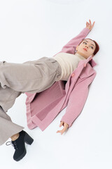 Asian woman poses lying on the floor in a pink coat, beige turtleneck, gray pants. Warm fashionable autumn clothes. White background.