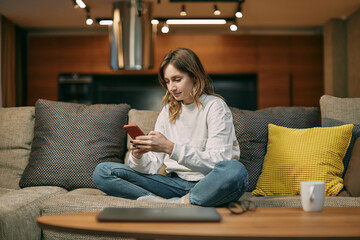 Young woman with phone sitting on sofa in living room, messaging in social media, shopping online,...