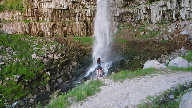 Girl standing in front of waterfall taking pictures. Water falling in slow motion. Drone slowly panning in.