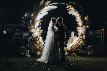 A stylish groom and a beautiful bride in a long dress are standing, hugging at night near a...