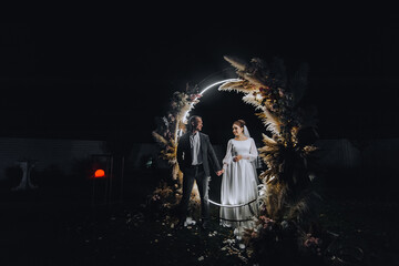 A stylish groom and a beautiful bride in a long dress stand holding hands, imitating a mirror, at night near a luminous reed arch decorated with lamps and garlands.