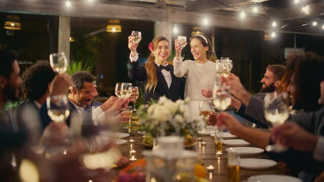 Big Dinner Party with a Crowd of Multiethnic Diverse Friends Celebrating at a Restaurant. Beautiful Happy Queer Hosts Propose a Toast and Raise Wine Glasses while Sitting at a Table in the Evening.